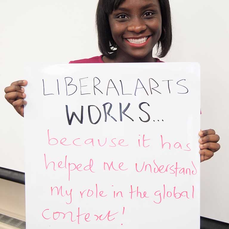A female student holds up a handwritten sign that reads ‘Liberal Arts Works...because it has helped me understand my role in the global context!’