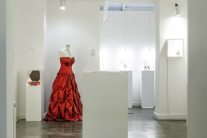 A photo from a Museum of Broken Relationships exhibit that shows a long, red strapless gown on a mannequin with four white pedestals near it.   