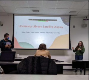 A picture of a classroom in which two students stand  on either side of a projector screen.