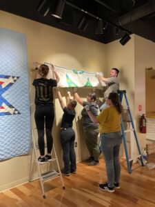 A group of people, some standing on ladders, hold onto a quilt as they remove it from the wall of a gallery.