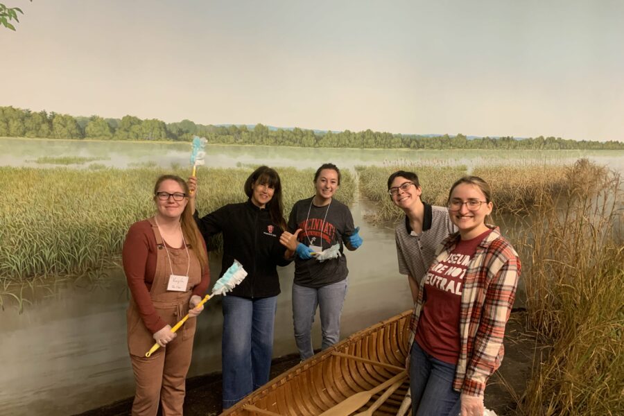 5 people standing in a museum display with a canoe smile while holding dusting tools.