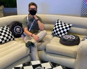 Syd Overtoom, a person with short brown hair, glasses, and wearing a face mask is sitting in the middle of a blow up white couch. This couch has 3 sections and has 3 checkered flag print patterned pillows and two inflatable pillows that look like car tires. There is the edge of a table directly in front of Syd with a checkered flag pattern as well. Syd is wearing a green button up shirt, white pants, and dark green shoes.